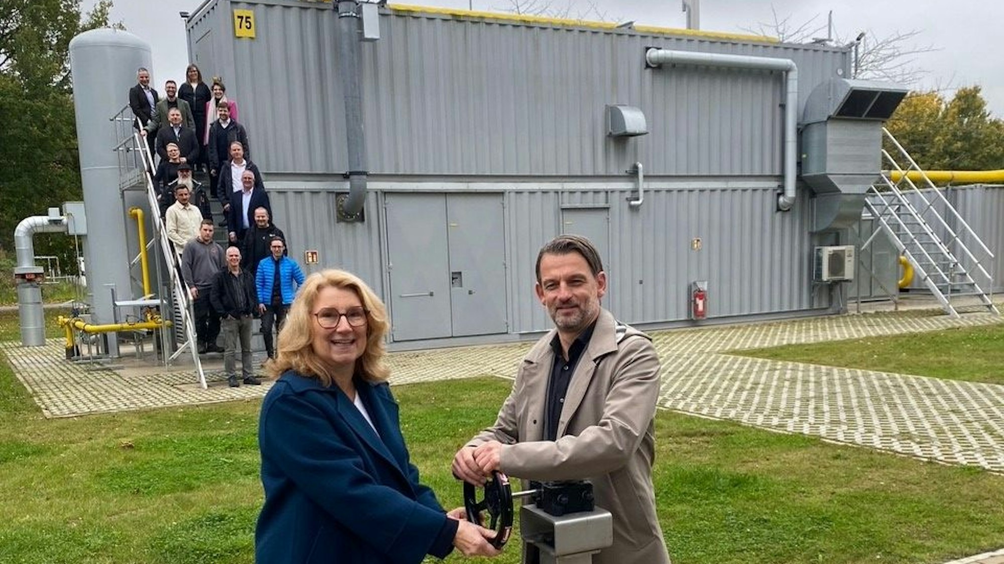 Steb-Vorständin Ulrike Franzke und Umweltdezernent William Wolfgramm nehmen die Biogasaufbereitungsanlage symbolisch in Betrieb; im Hintergrund die Projektbeteiligten auf der Treppe der Anlage.