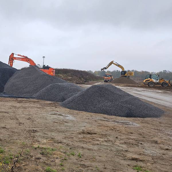 Auf einer Baustelle liegen Rohre und Schotterhaufen, im Hintergrund arbeiten Bagger.