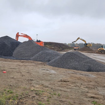 Auf einer Baustelle liegen Rohre und Schotterhaufen, im Hintergrund arbeiten Bagger.