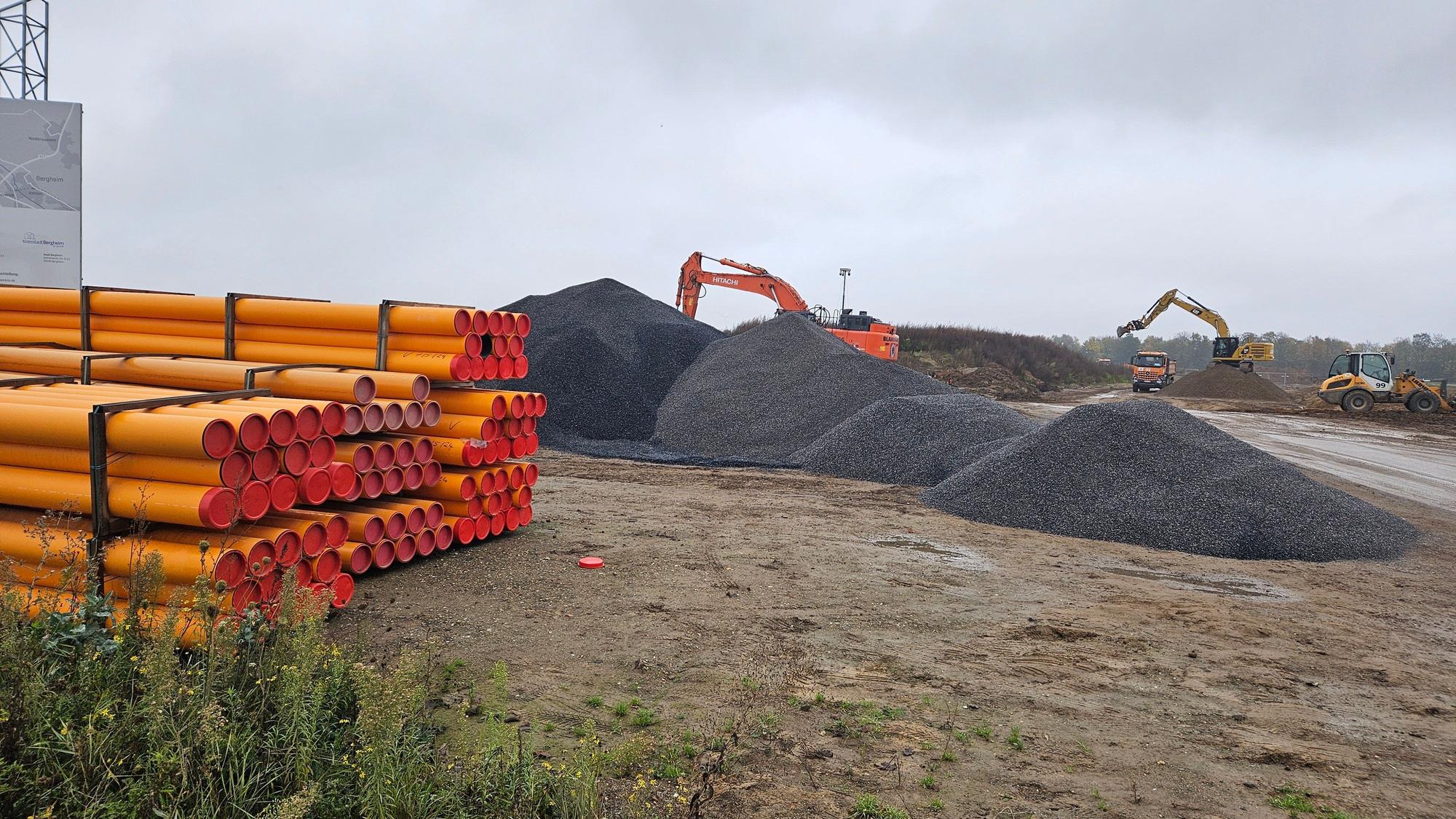 Auf einer Baustelle liegen Rohre und Schotterhaufen, im Hintergrund arbeiten Bagger.