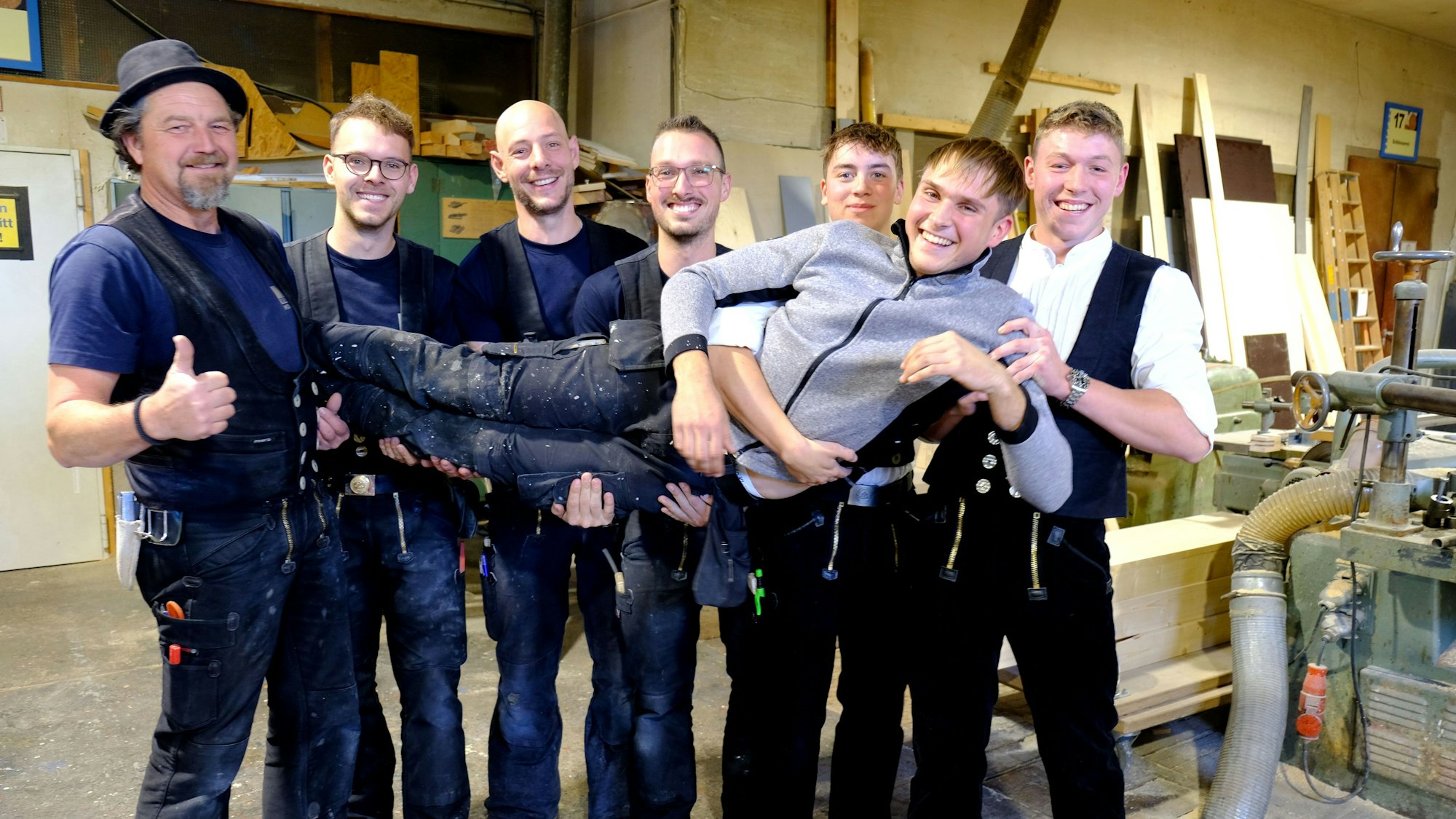 Patrick van Kann (liegend) wird von seinen Kollegen der Firma Alois und Leo Völler Holzprodukte aus Sistig auf Händen getragen. Die Gruppe steht vor Geräten und Material einer Holzwerkstatt.