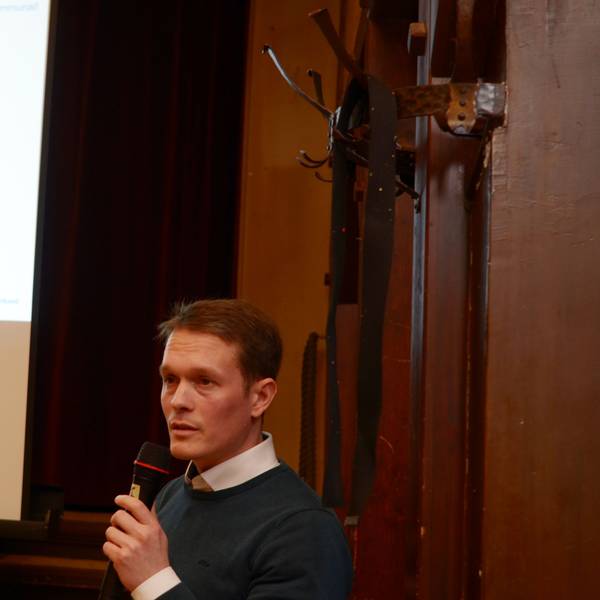 Ein Mann mit einem Mikrofon in der Hand hält einen Vortrag. Im Hintergrund ist eine Präsentation zu erkennen.