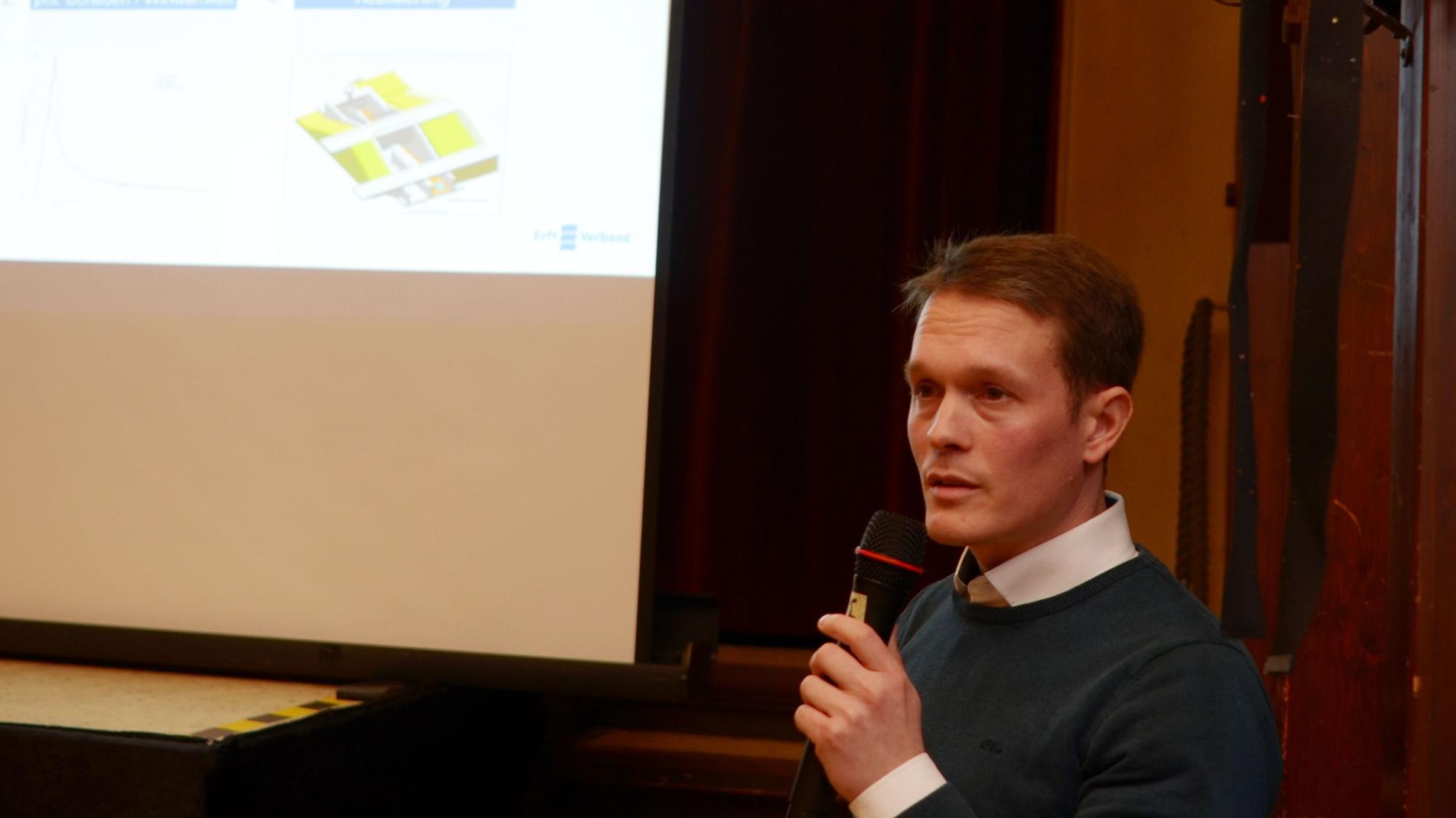 Ein Mann mit einem Mikrofon in der Hand hält einen Vortrag. Im Hintergrund ist eine Präsentation zu erkennen.