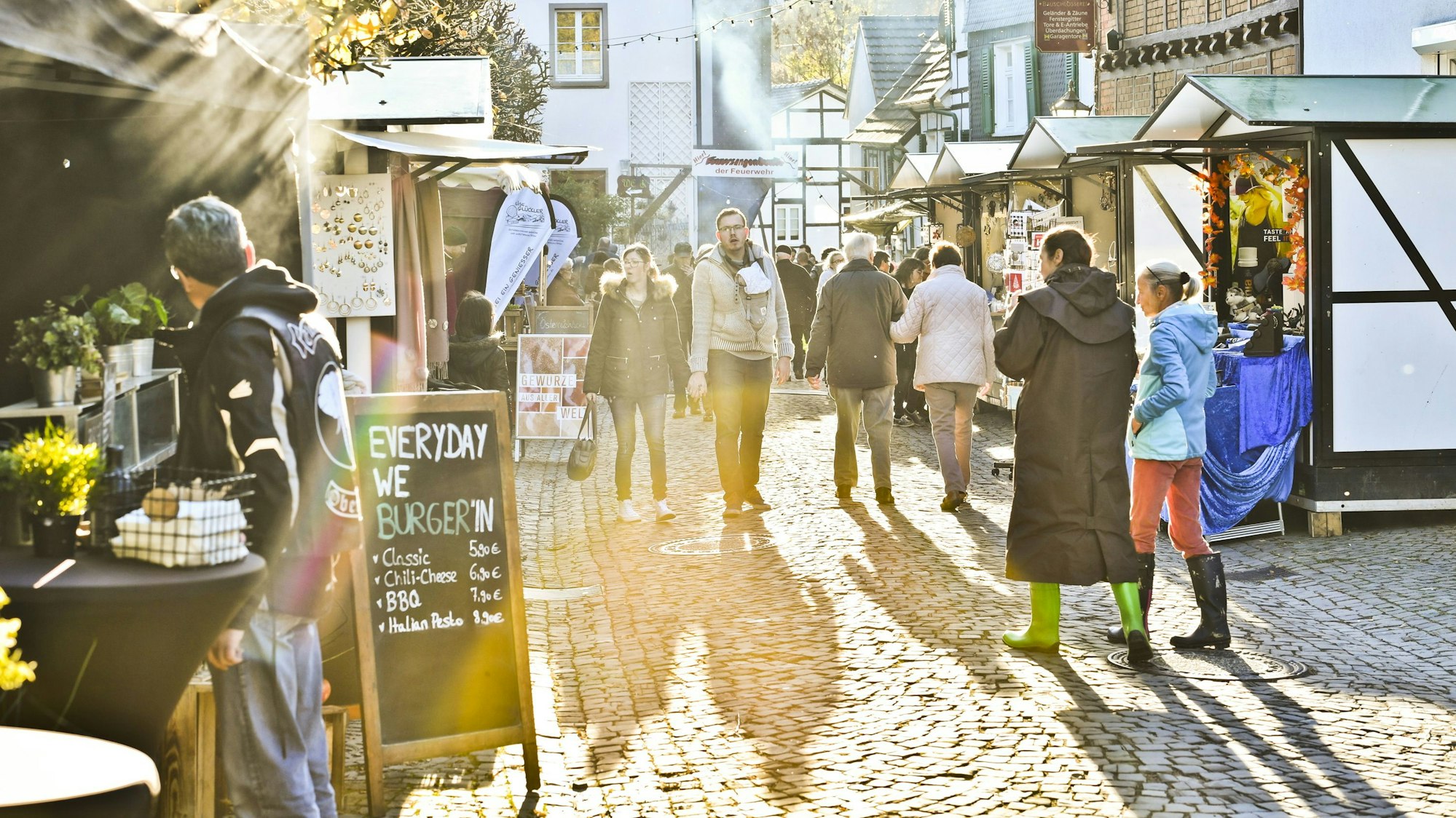 Blick in eine von Buden gesäumte Straße mit Fachwerkhäusern und Kopfsteinpflaster, Menschen schlendern an Verkaufsständen entlang. Sonnenstrahlen tauchen die Szene in warmes Licht.