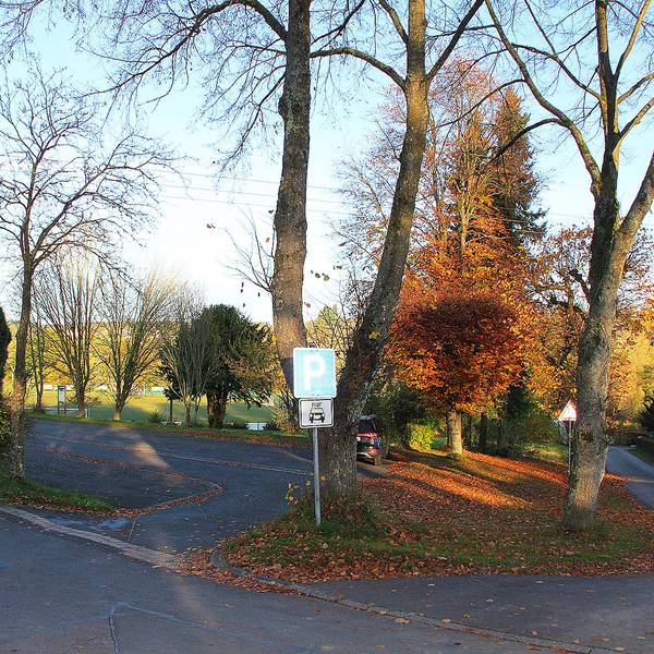 Nahe des Sportplatzes in Dahlem-Schmidtheim ist ein kleiner Parkplatz, der in einem schlechten Zustand ist. Rund um den asphaltierten Platz sind Bäume zu sehen.
