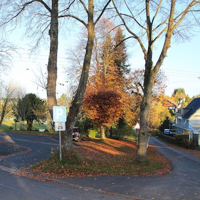 Nahe des Sportplatzes in Dahlem-Schmidtheim ist ein kleiner Parkplatz, der in einem schlechten Zustand ist. Rund um den asphaltierten Platz sind Bäume zu sehen.