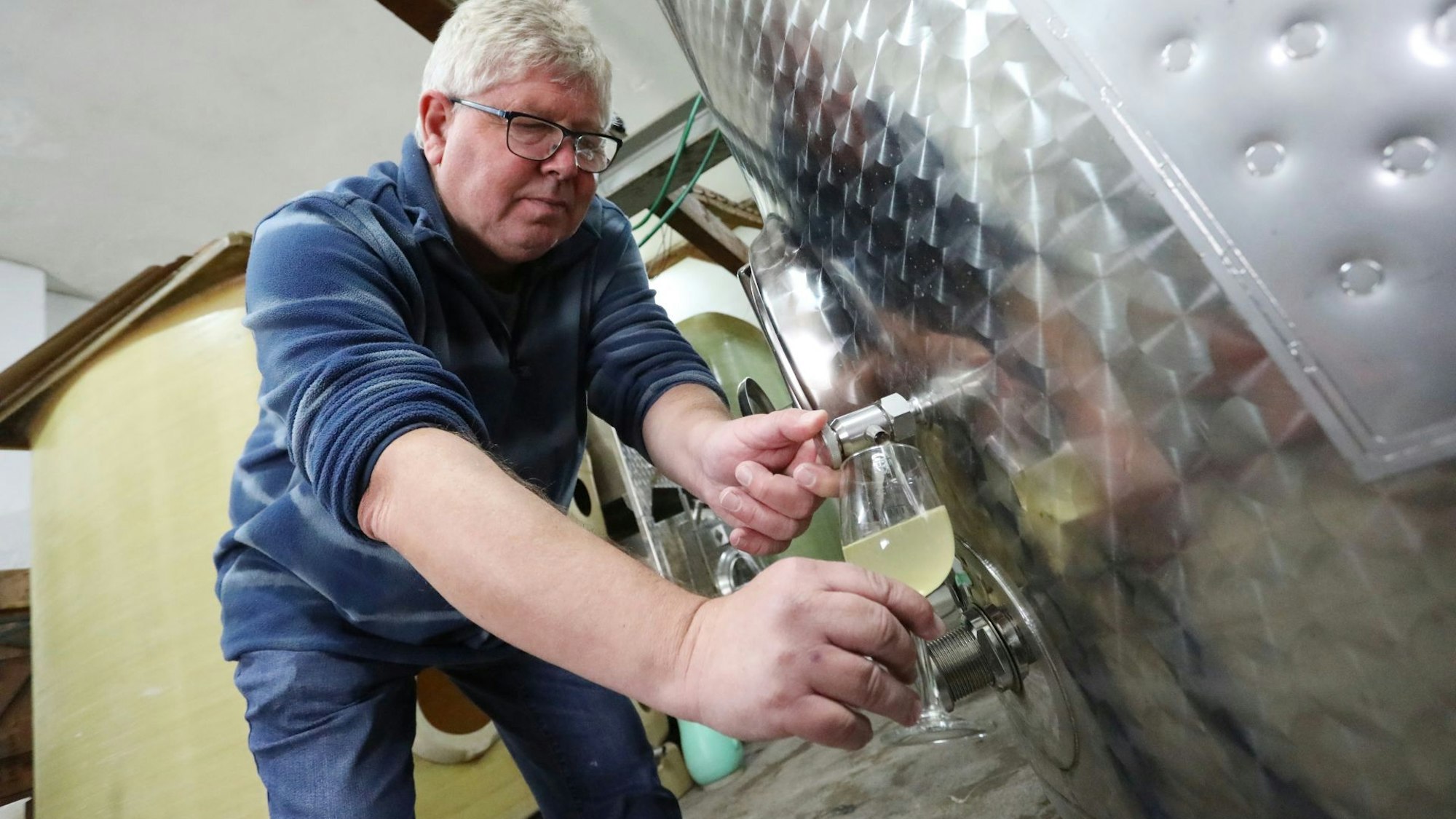 Winzer Bernd Blöser mit neuem Wein der Ernte 2024, der noch im Fass reifen kann. Er zapft gerade ein Glas aus dem Fass ab. Der junge Wein ist noch trüb.