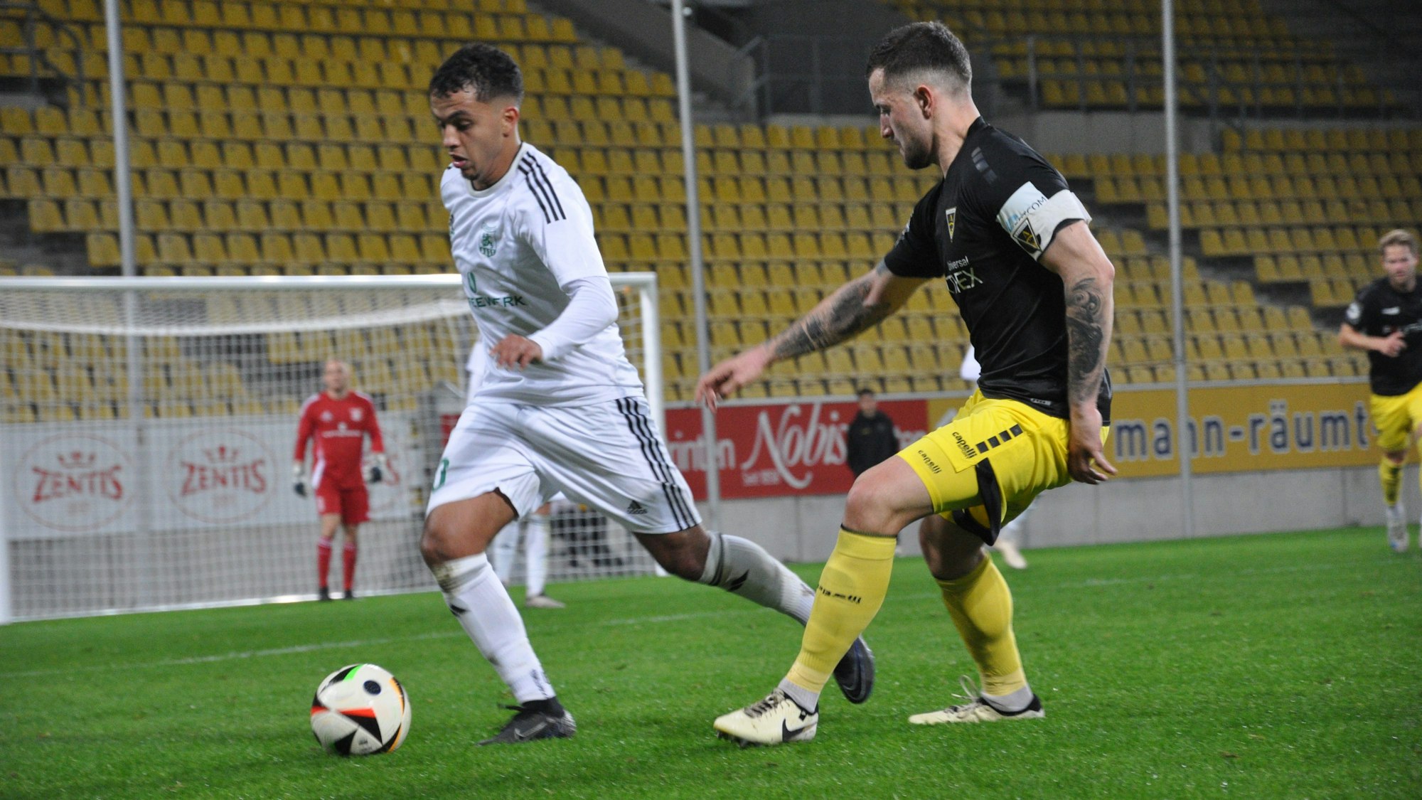 Das Foto zeigt einen Fußballspieler in einem weißen Trikot am Ball, sein Gegner trägt ein schwarz-gelbes Trikot.