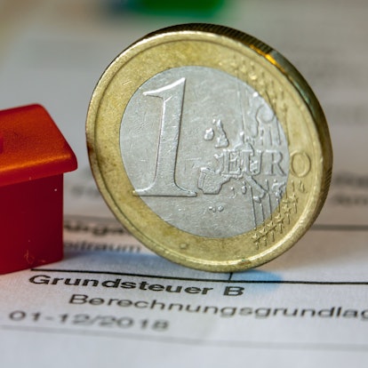 Eine Euro-Geldmünze sowie ein Spielzeughaus stehen auf einem Abgabenbescheid für die Entrichtung der Grundsteuer.