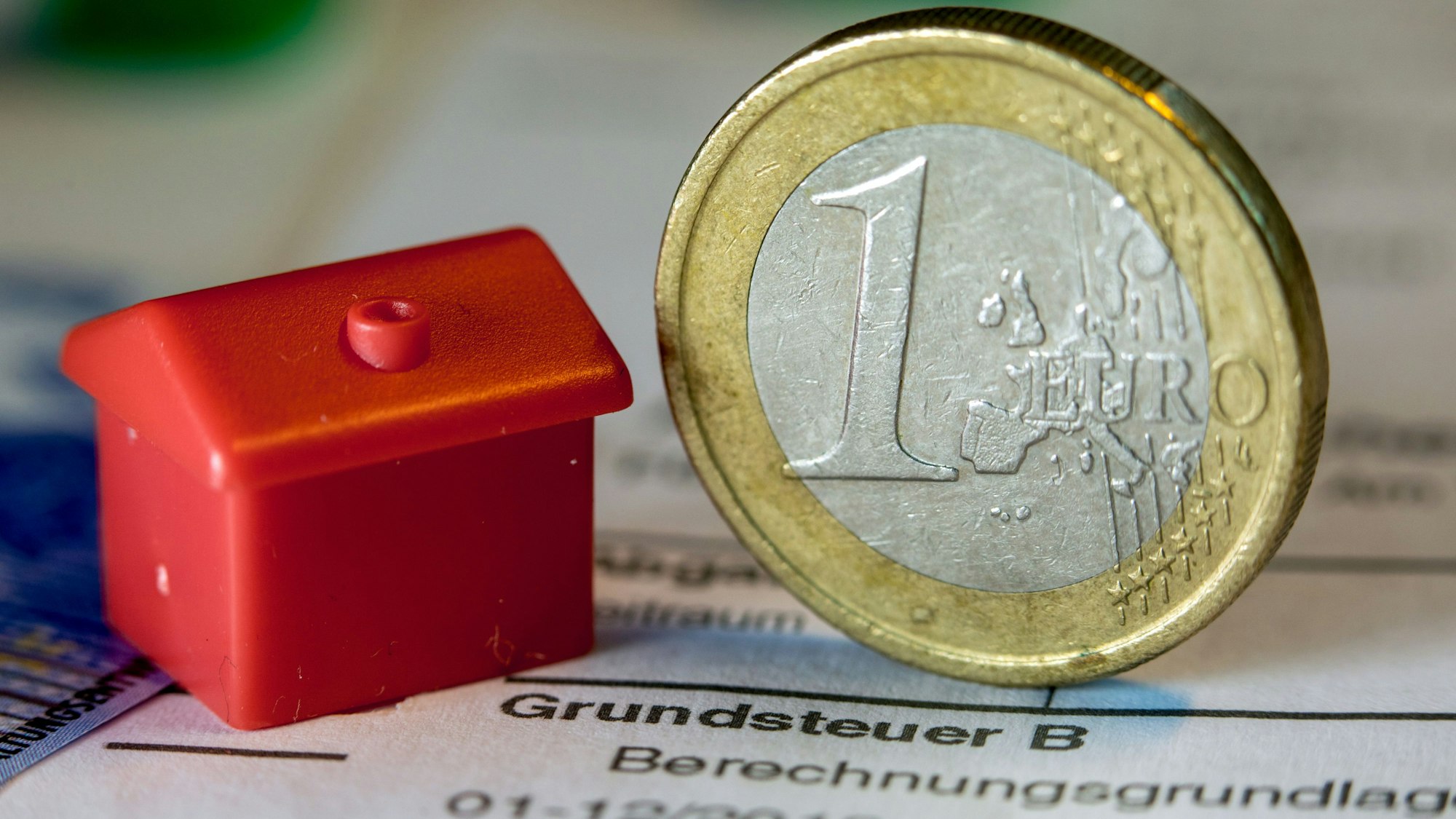 Eine Euro-Geldmünze sowie ein Spielzeughaus stehen auf einem Abgabenbescheid für die Entrichtung der Grundsteuer.