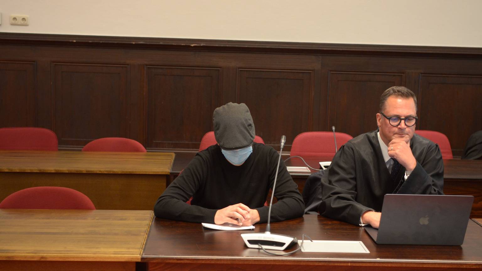 Einer der Angeklagten sitzt mit geneigtem Kopf, Schutzmaske und Kappe neben seinem Verteidiger.