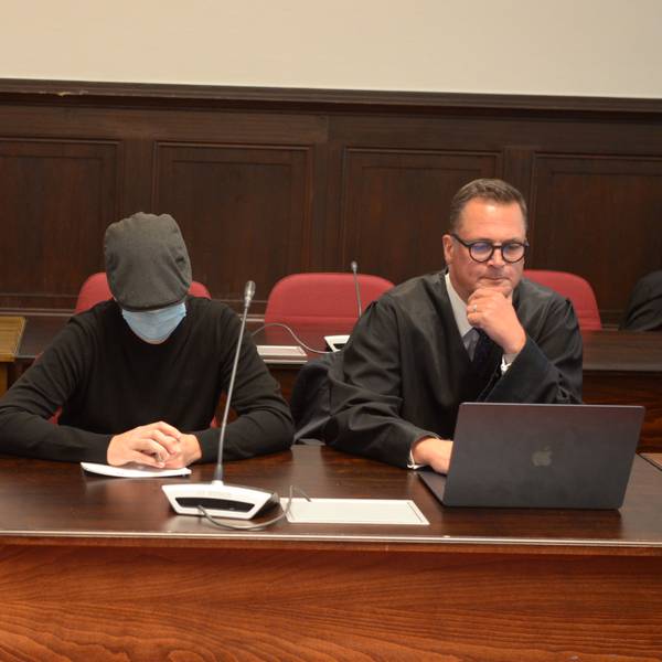 Einer der Angeklagten sitzt mit geneigtem Kopf, Schutzmaske und Kappe neben seinem Verteidiger.