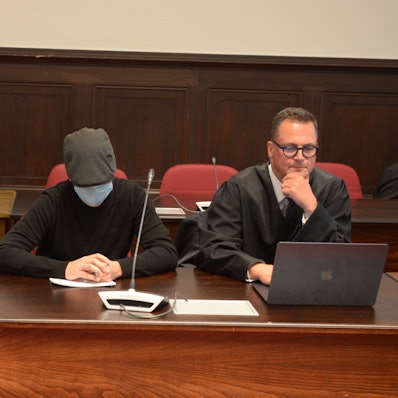 Einer der Angeklagten sitzt mit geneigtem Kopf, Schutzmaske und Kappe neben seinem Verteidiger.