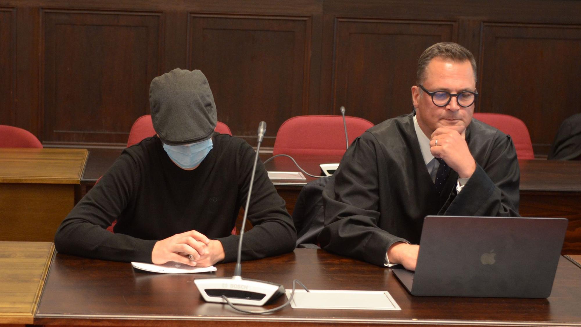 Einer der Angeklagten sitzt mit geneigtem Kopf, Schutzmaske und Kappe neben seinem Verteidiger.