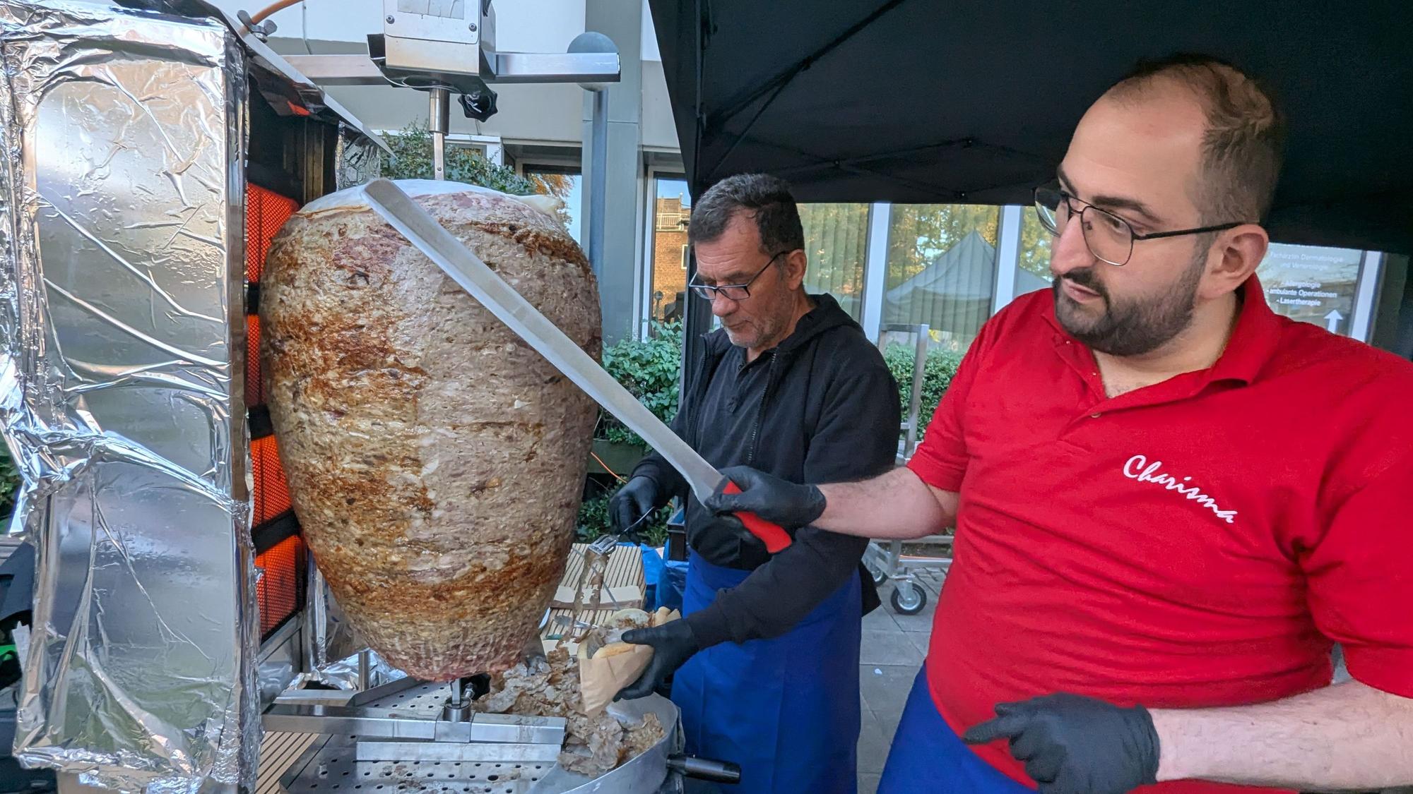Das Foto zeigt zwei Männer, einer davon schneidet mit einem großen Messer Fleisch von einem Dönerspieß.