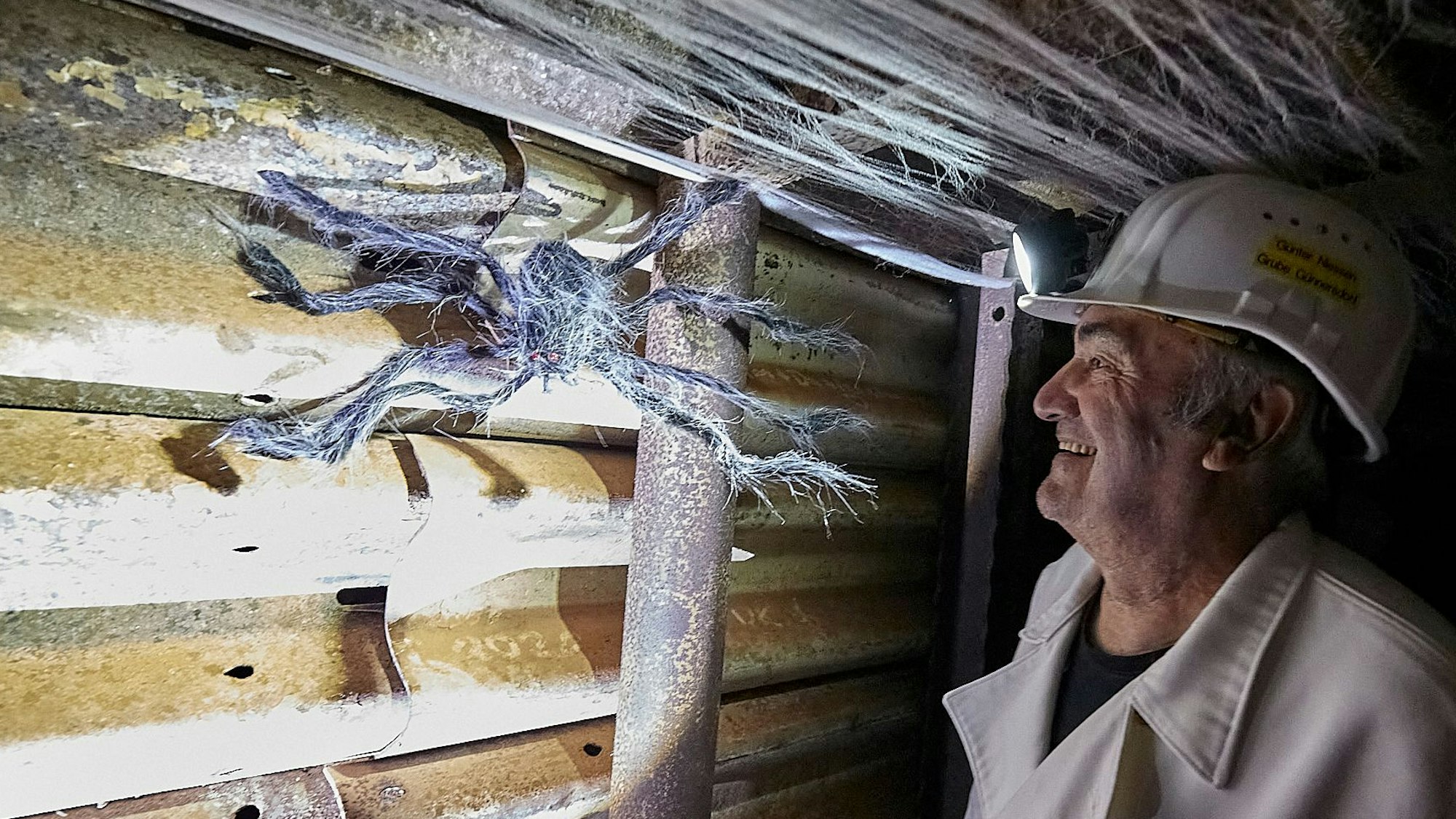 Günter Nießen trägt die weiße Kleidung eines Bergmanns und betrachtet Besucherbergwerk Grube Günnersdorf eine große Spinnenfigur, die als Halloween-Dekoration an der Wand angebracht ist.