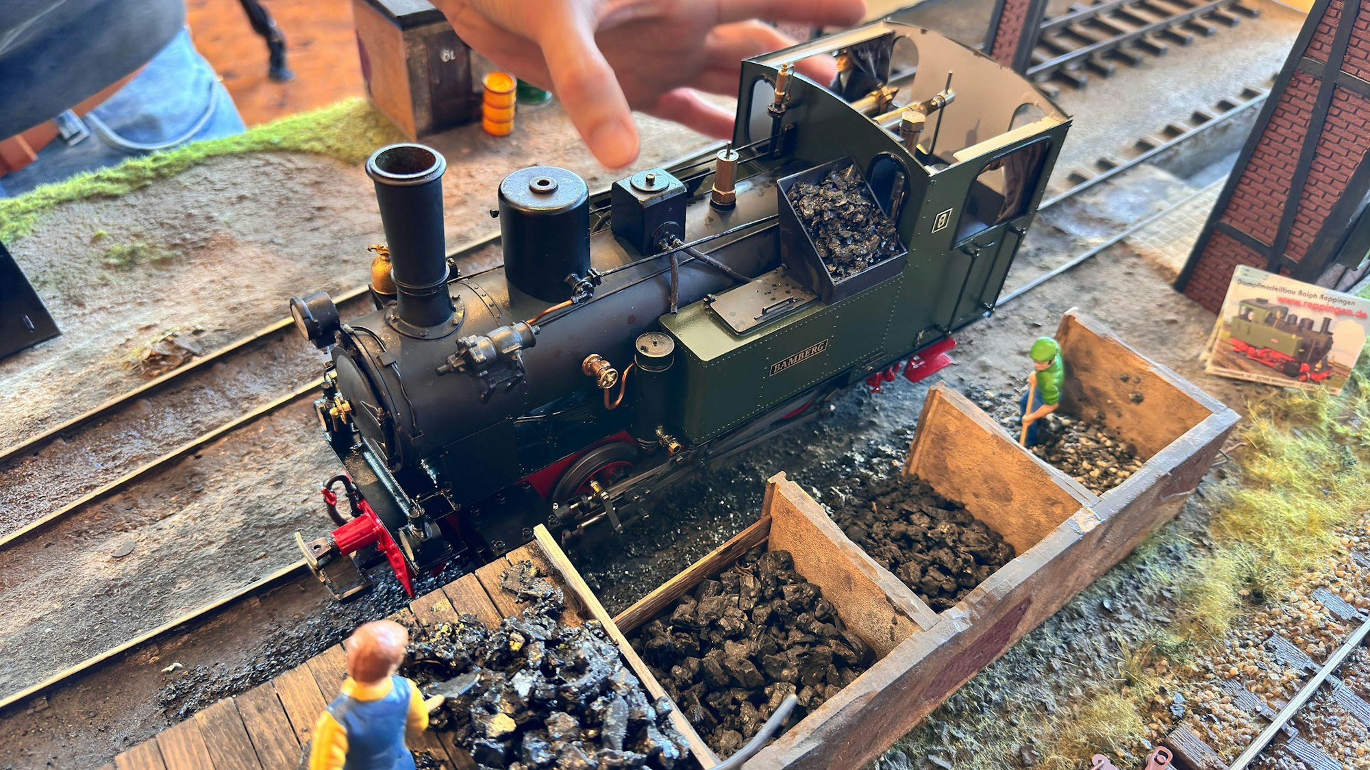 Neben der klassischen schwarzen Lok warten kleine Figuren an einem Kohlelager.