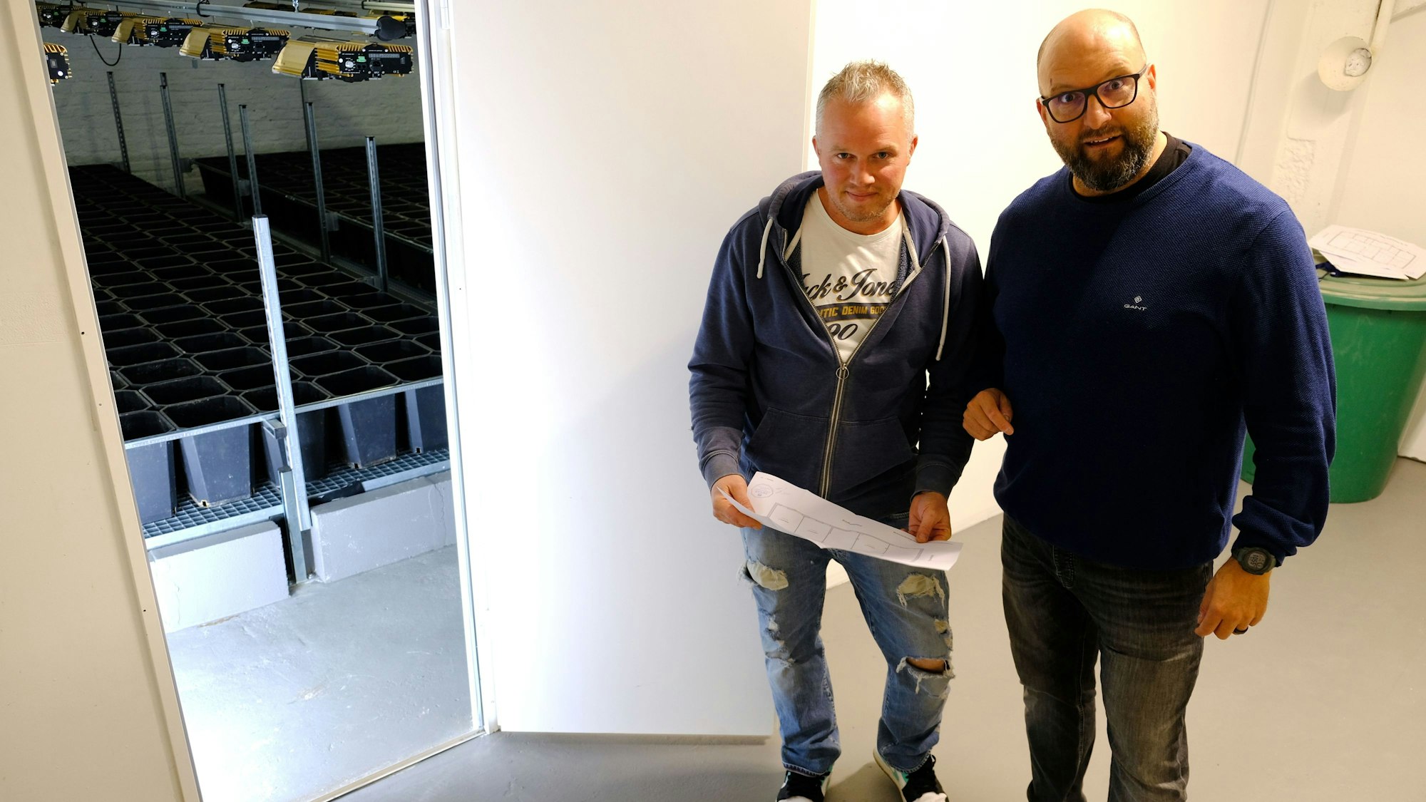 Zwei Männer stehen vor einer offenen Tür, die zu einem Raum führt, in dem die Ausrüstung für den Anbau von Cannabis-Pflanzen steht.
