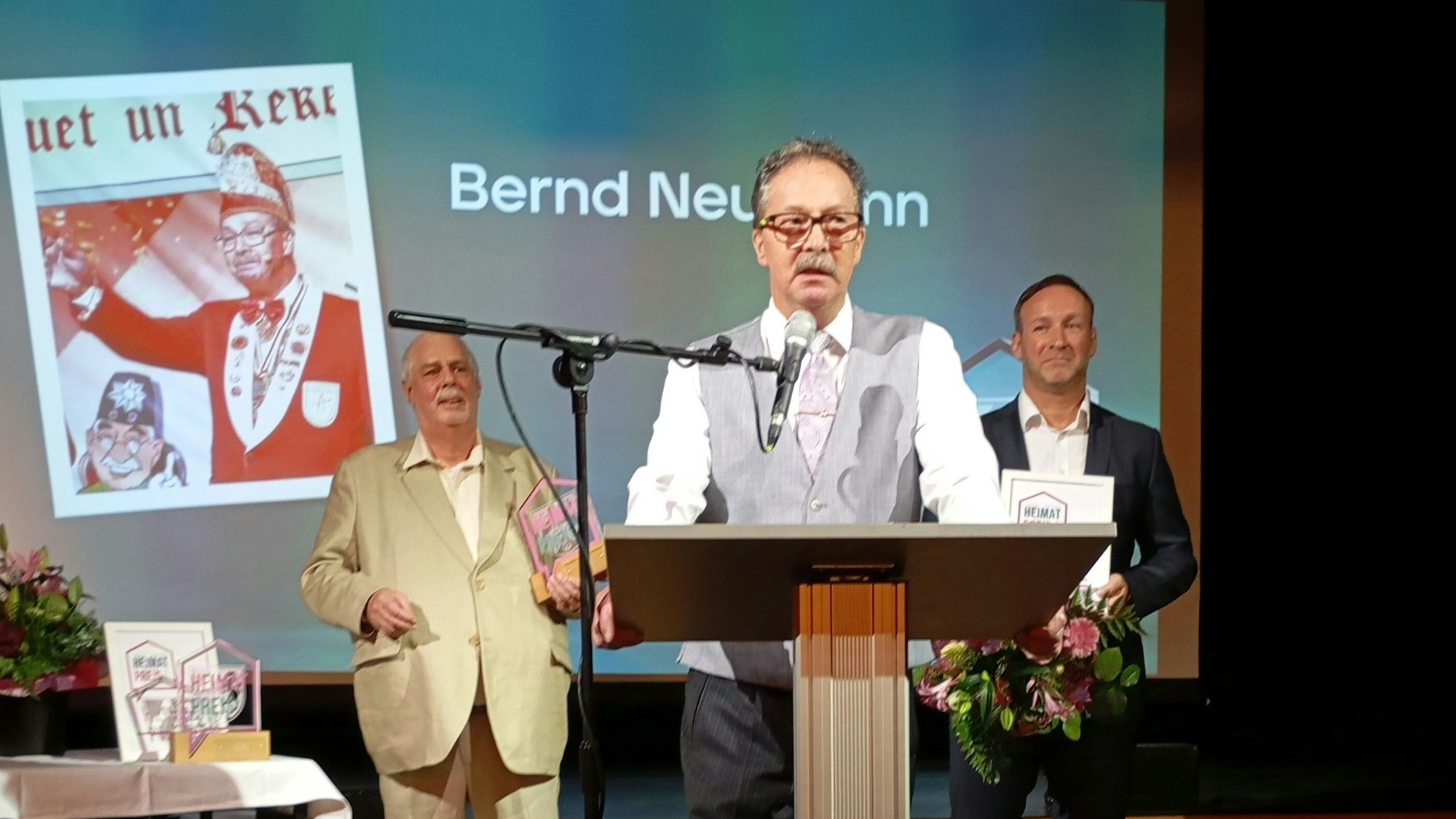 Bernd Neumann trägt eine graue Weste und ein weißes Hemd. Er steht an einem Pult und spricht in ein Mikrofon.