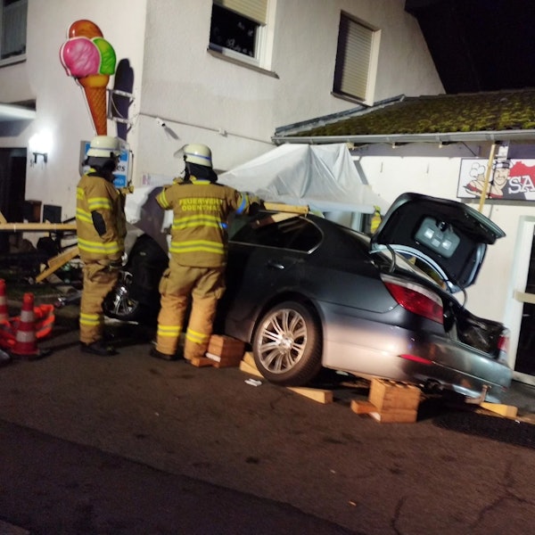 Zwei Feuerwehrleute stehen neben einem Unfallauto vor einem Gebäude mit einem Eiscafé-Schild.