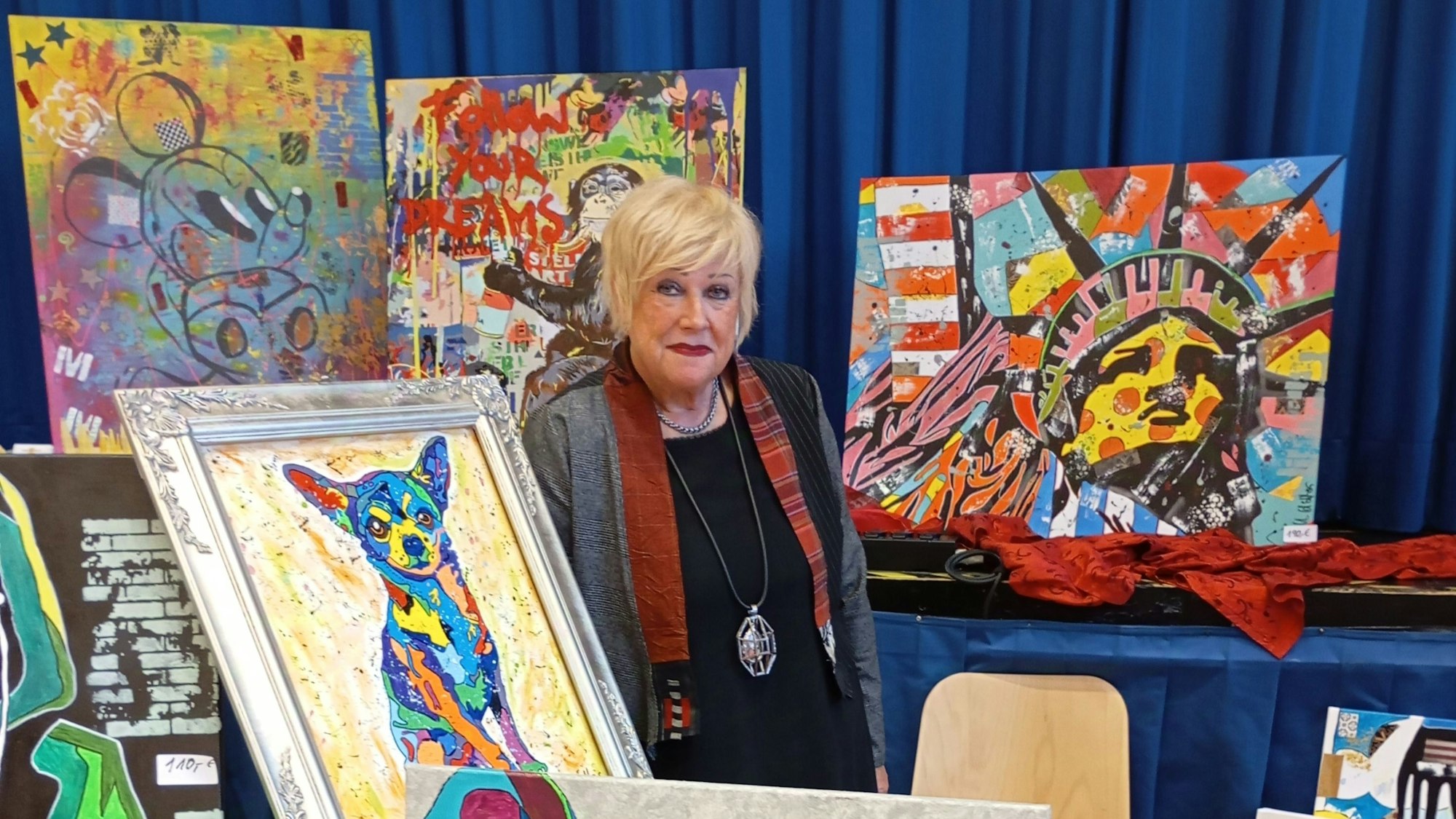 Ulrike Höllers steht umringt von ihren bunten Bildern, die Motive wie die Freiheitsstatue, einen Hund und Micky Maus zeigen.