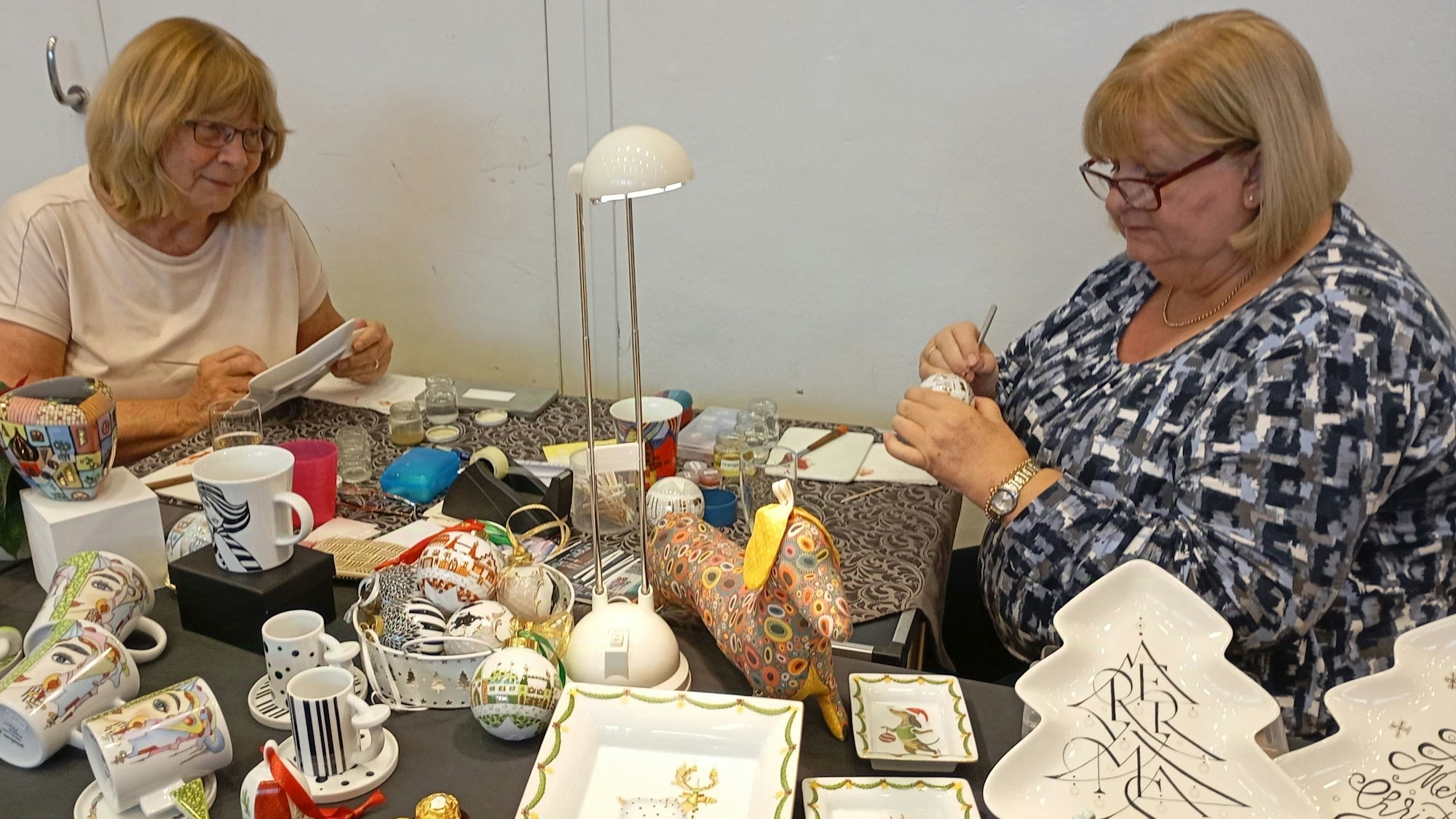 Während des Marktes bemalen die beiden Damen weiteres Porzellan. Ihre Stücke präsentieren sie auf einem Tisch.