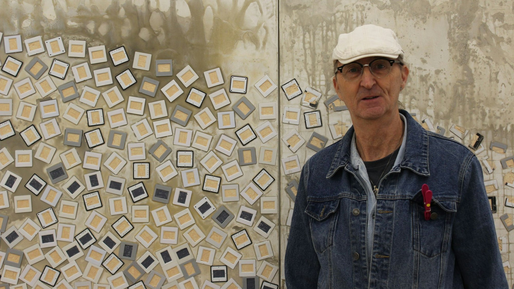 Bernhard Johannes Trier steht vor seinem großen Kunstwerk an der Wand. Er trägt eine Jeansjacke und eine weiße Mütze.