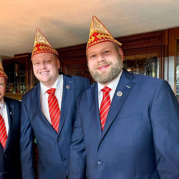 Drei Männer in identischen blauen Anzügen mit rot-weiß-gestreiften Krawatten und Schiffchen lachen in die Kamera.