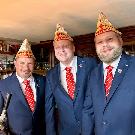 Drei Männer in identischen blauen Anzügen mit rot-weiß-gestreiften Krawatten und Schiffchen lachen in die Kamera.