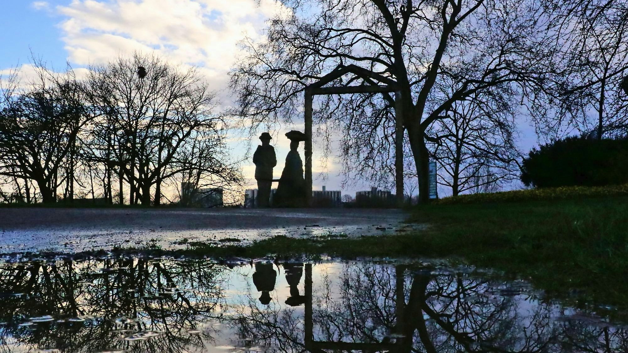 In einer Pfütze spiegeln sich Bäume und zwei lebensgroße Figuren, sowie Bäume.
