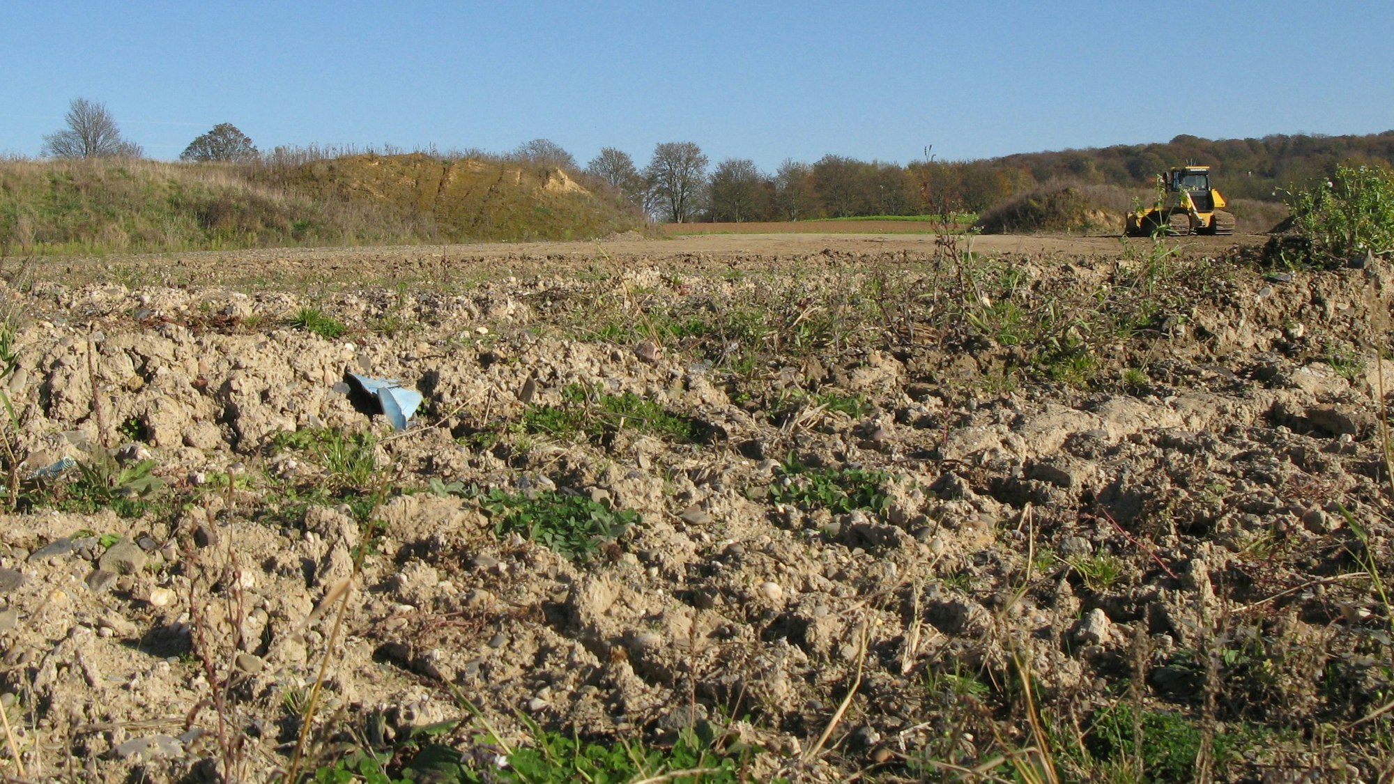 Blick auf die Erddeponie in Hennef-Petershohn mit einem planierten Boden im Vordergrund und einem gelben Bagger und Erdhügeln im Hintergrund.
