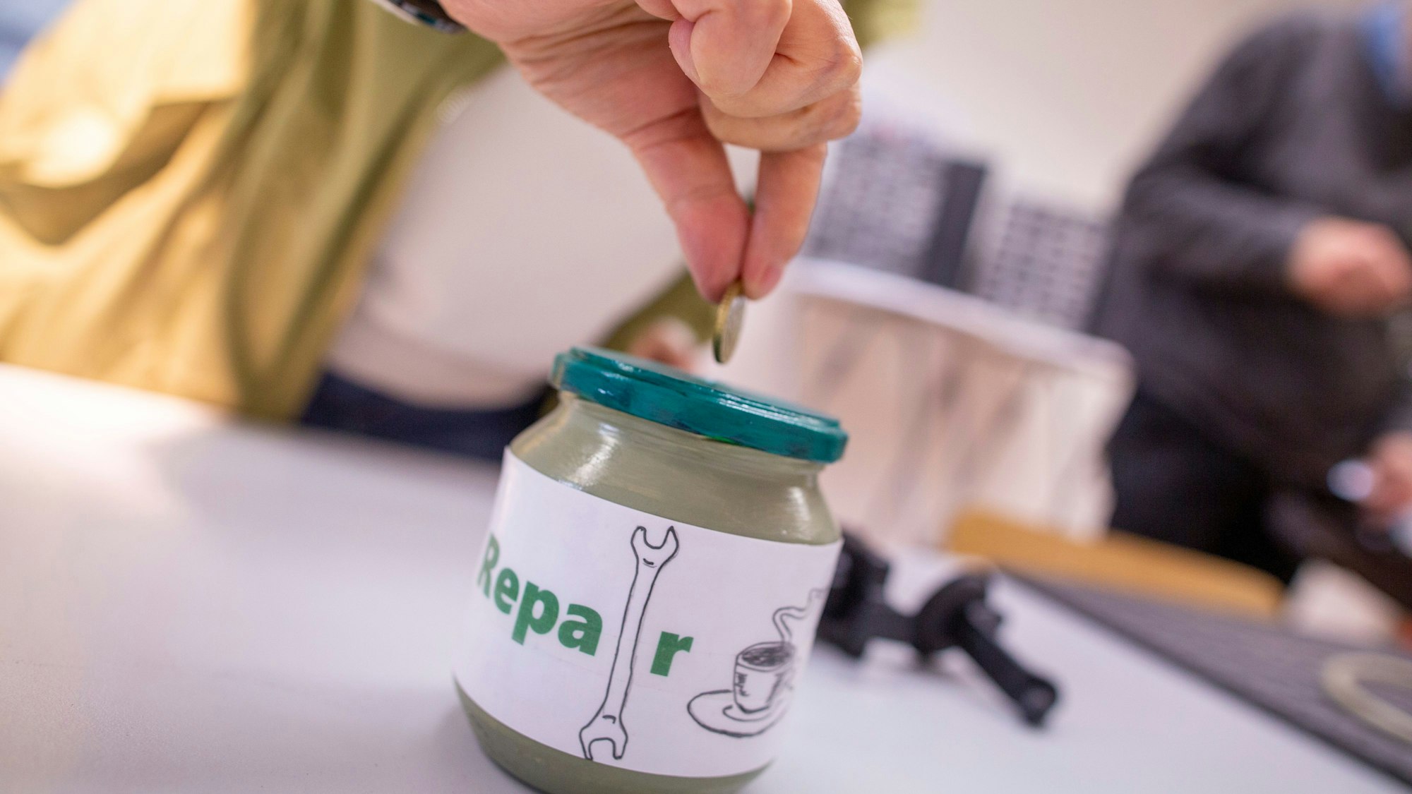 Eine Hand steckt eine Münze in die Kaffeekasse des Repair-Cafés.