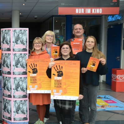 Eine Gruppe von sechs Frauen und zwei Männern hat sich zum Gruppenfoto aufgestellt. Sie tragen orange Plakate mit einer schwarzen zum Stopp gespreizten Handfläche.