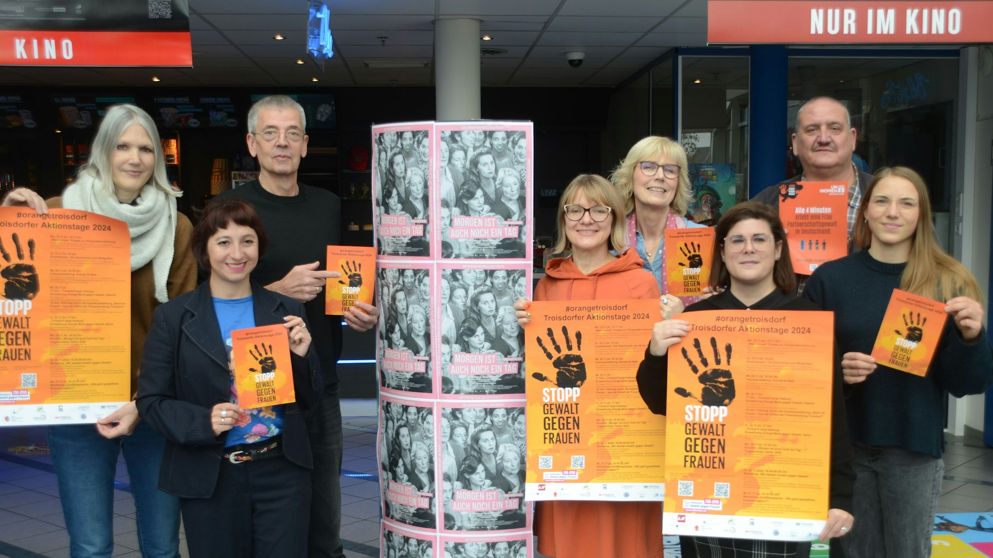 Eine Gruppe von sechs Frauen und zwei Männern hat sich zum Gruppenfoto aufgestellt. Sie tragen orange Plakate mit einer schwarzen zum Stopp gespreizten Handfläche.