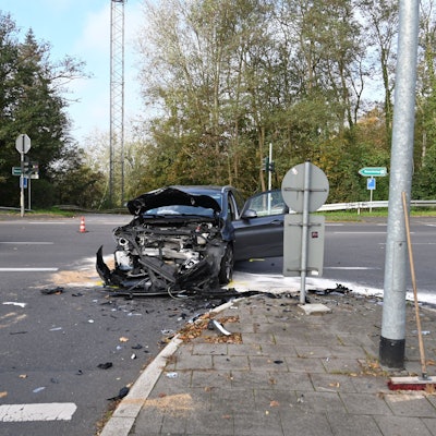 Ein an der Front vollkommen zerstörtes Auto steht auf einer Straßenkreuzung. Links im Hintergrund sind Teile eines Feuerwehrautos zu sehen. Auf der Straße liegen Splitter verteilt.