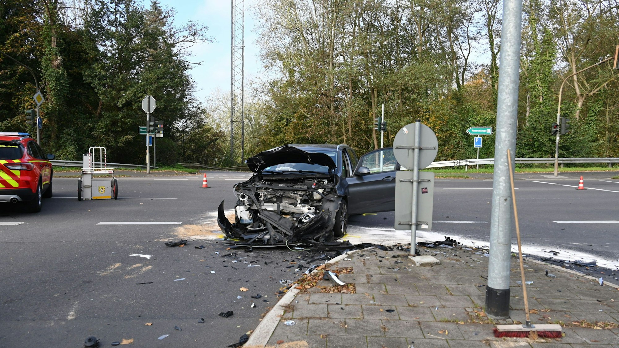 Ein an der Front vollkommen zerstörtes Auto steht auf einer Straßenkreuzung. Links im Hintergrund sind Teile eines Feuerwehrautos zu sehen. Auf der Straße liegen Splitter verteilt.