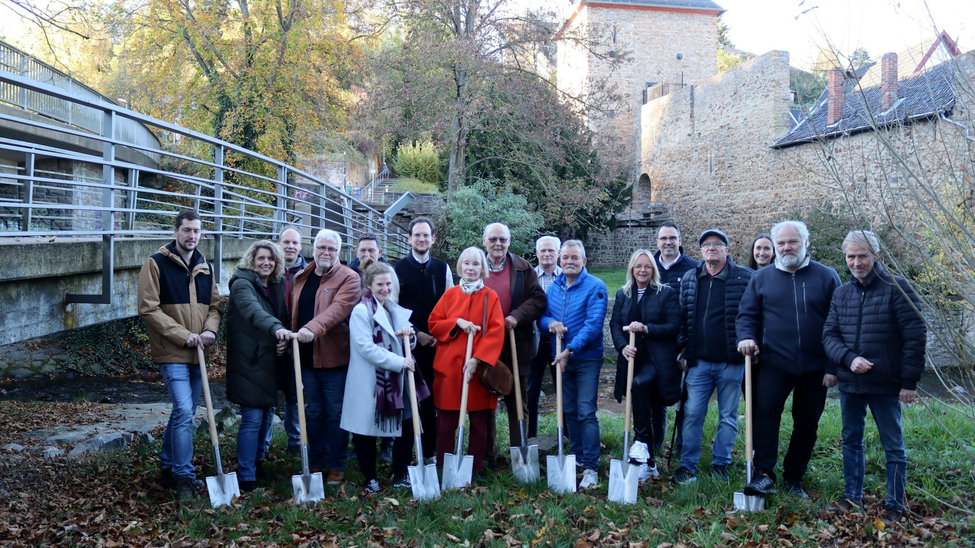 In der Nähe der Erft, im Hintergrund das Heisterbacher Tor in Bad Münstereifel, stehen Menschen mit einem Spaten in der Hand auf einer Wiese.