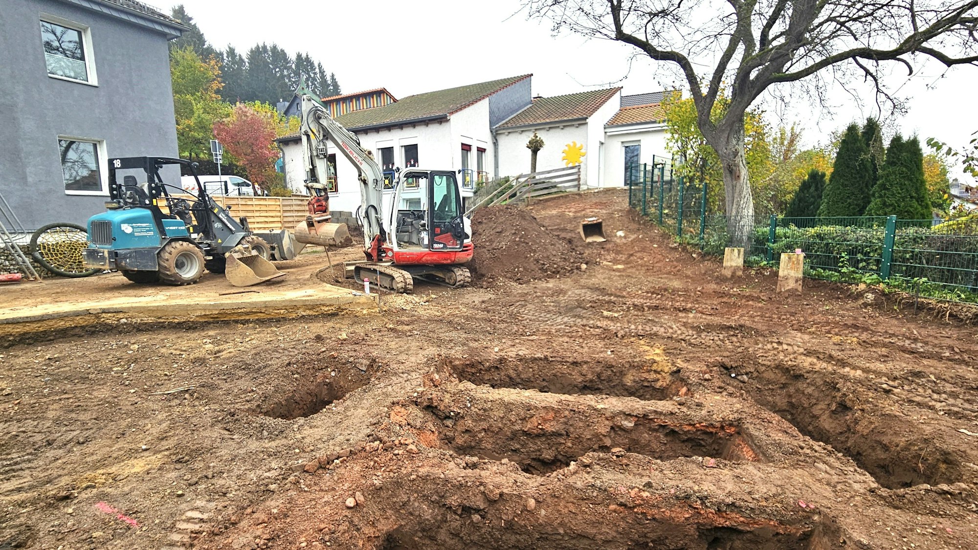 Ein Bagger und eine Raupe stehen auf einer Fläche, deren Boden abgetragen ist und die mehrere Löcher aufweist.