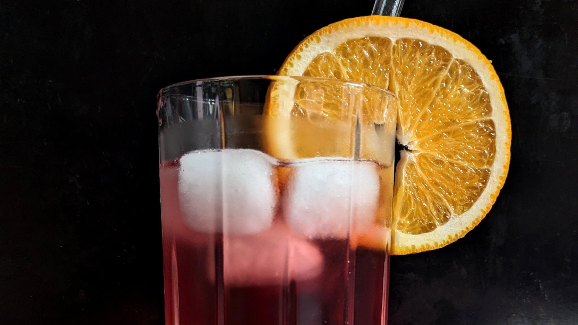 An einem Glas mit Eiswürfeln und dem roten Getränk hängt eine Orangenscheibe.