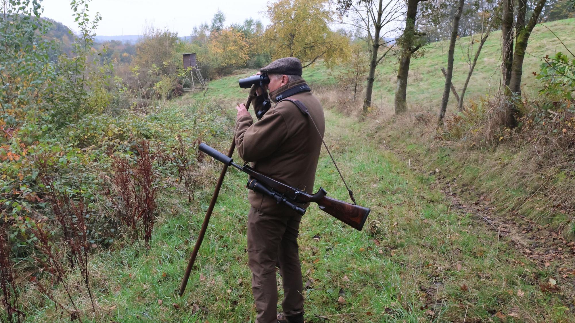 Ein Mann mit umgehängtem Gewehr schaut mit einem Fernglas in den Wald.