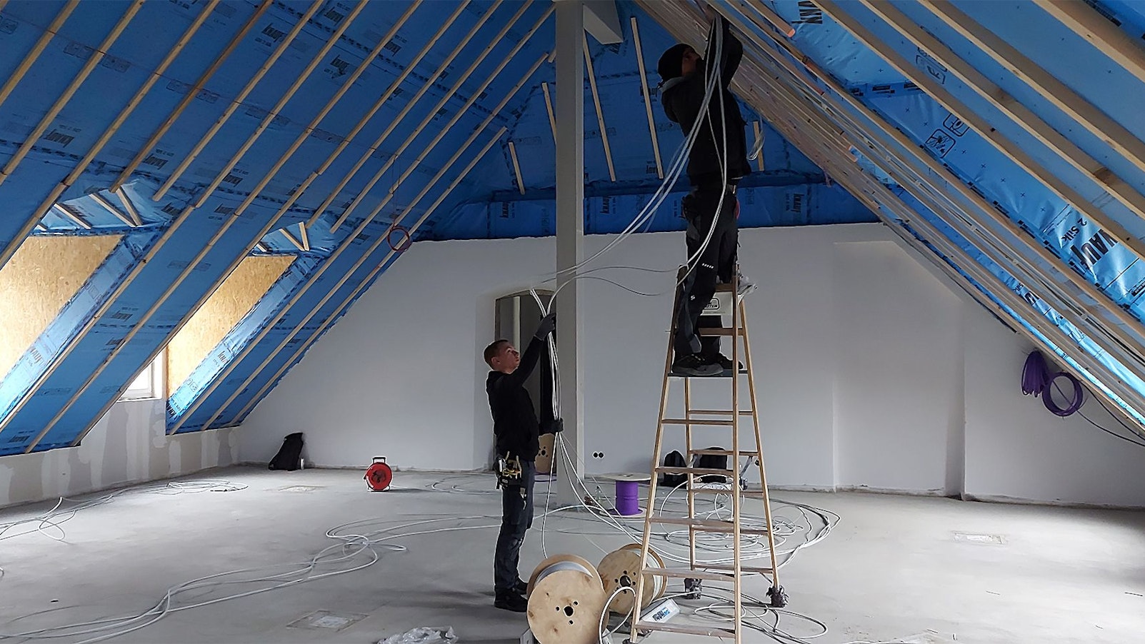Zwei Handwerker arbeiten im Dachgeschoss eines Hauses. Einer der Männer steht auf einer Leiter.