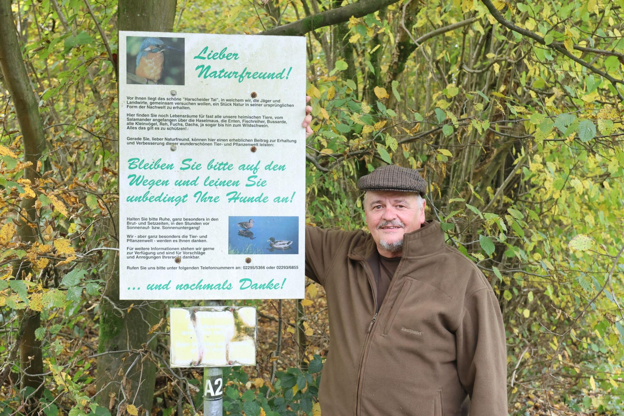 Jäger Steinhausen steht neben einem Schild am Wegesrand im Wald, er wirbt für Rücksicht auf die Natur und informiert Wanderer.
