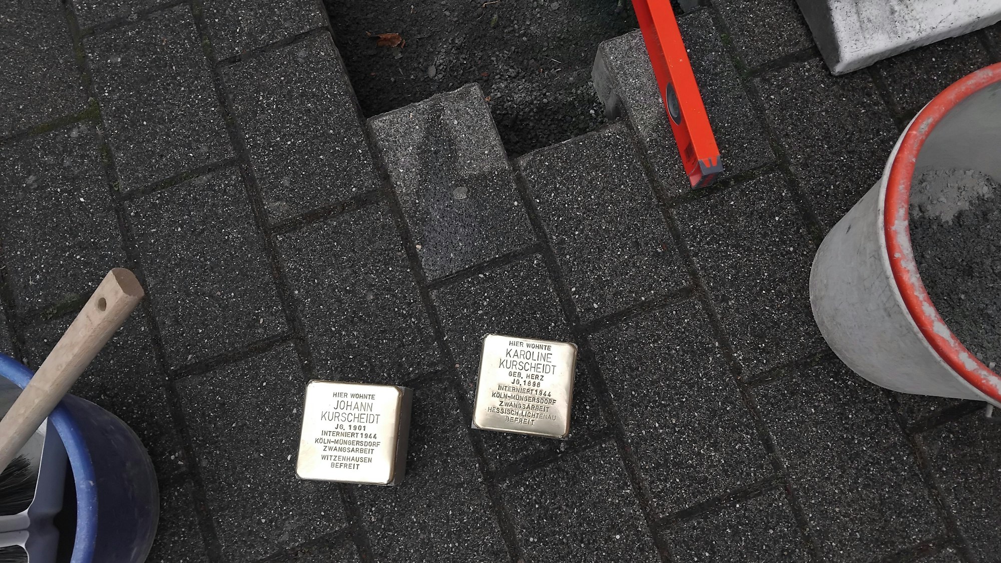 Zwei Stolpersteine mit den Namen Karoline Kurscheidt und Johann Kurscheidt liegen kurz vor ihrer Verlegung auf dem Bordstein, daneben sind Eimer und Werkzeuge zu sehen.