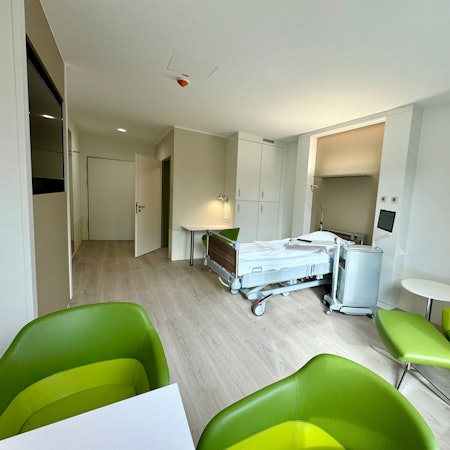 Auf dem Bild ist eines der Zimmer zu sehen, mit einem Krankenhausbett und grünen Sesseln.