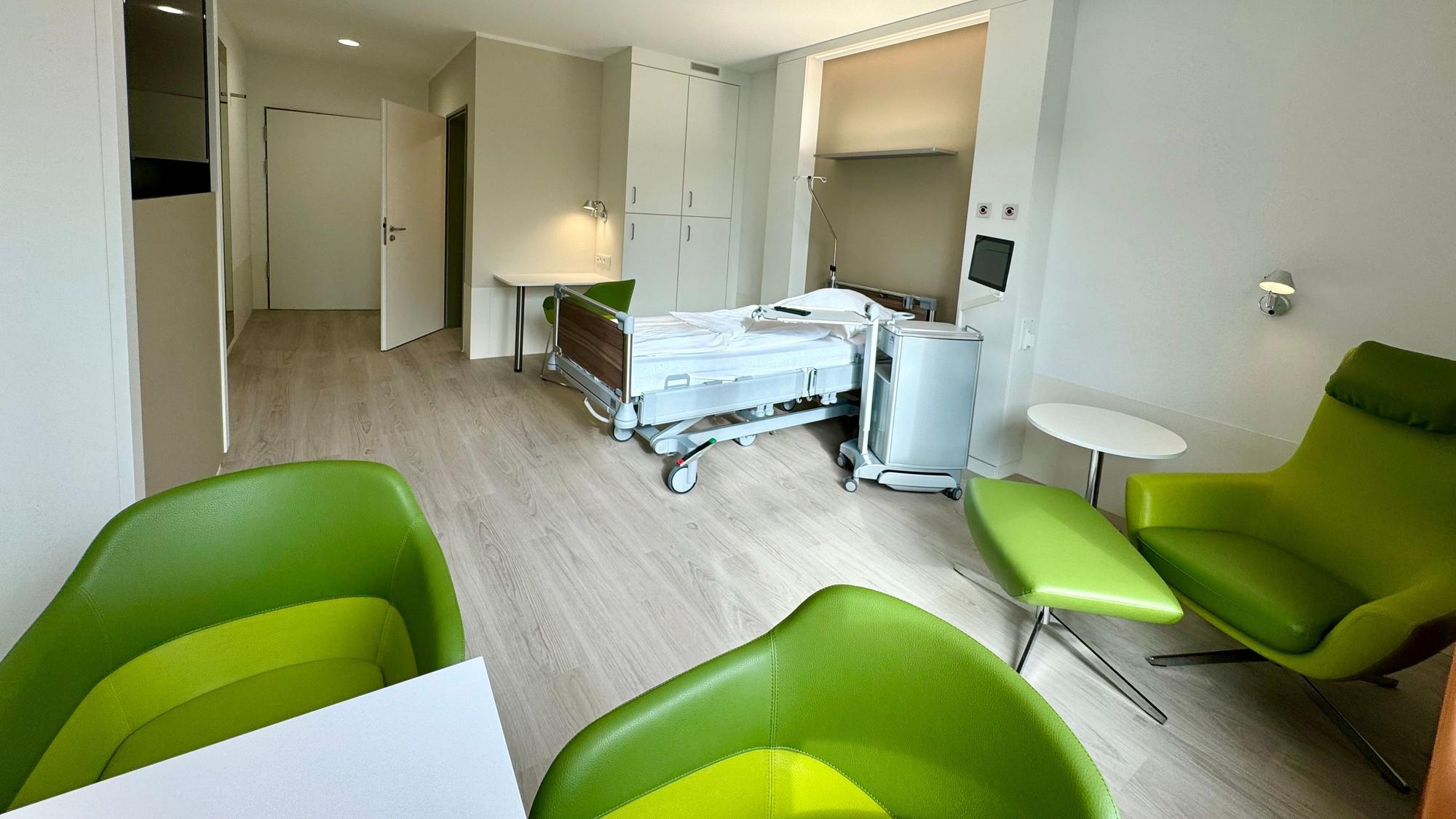 Auf dem Bild ist eines der Zimmer zu sehen, mit einem Krankenhausbett und grünen Sesseln.