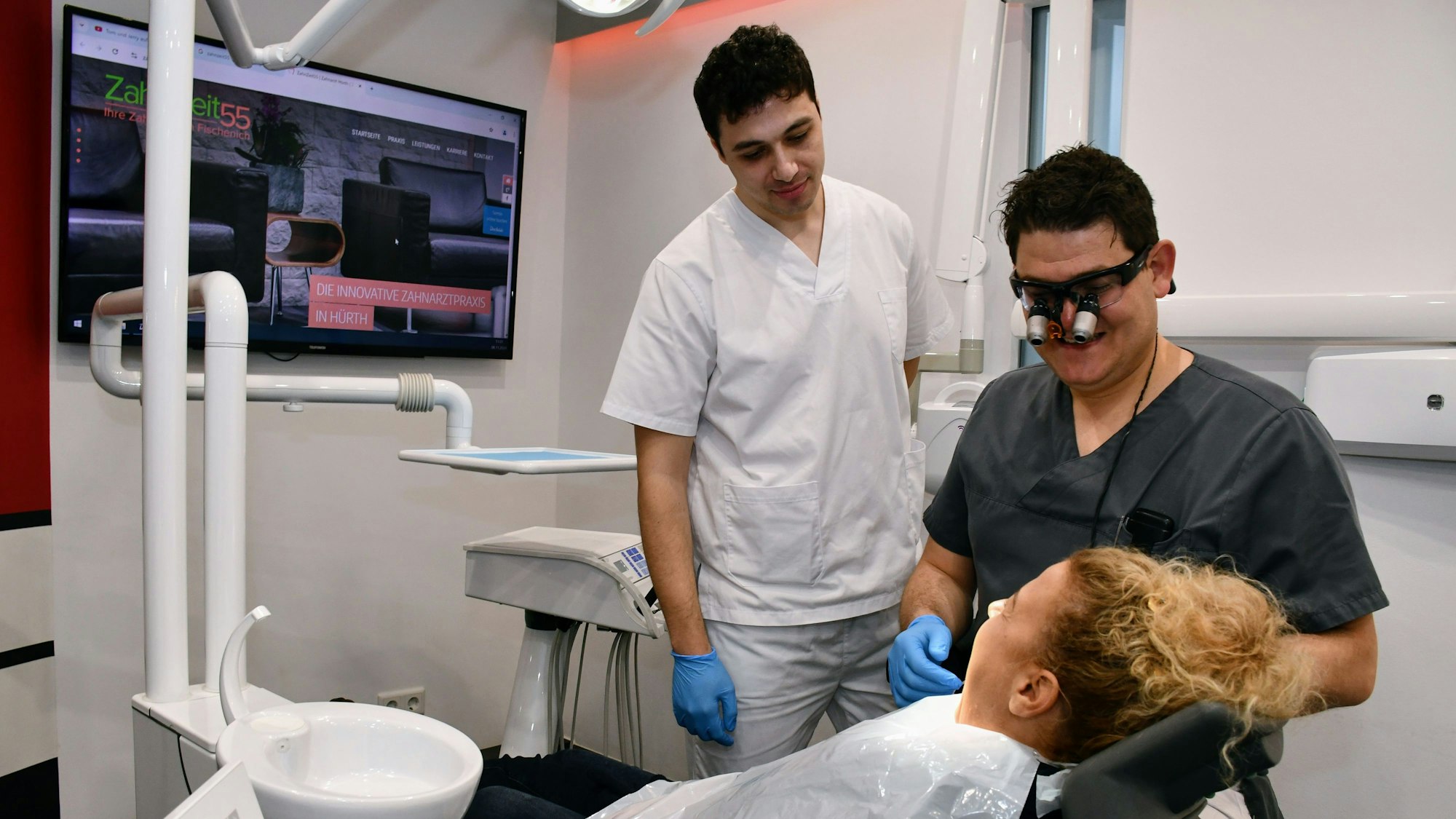Das Foto zeigt einen Auszubildenden im weißen Kittel und den Zahnarzt mit Lupenbrille vor dem Behandlungsstuhl, auf dem eine Frau liegt.