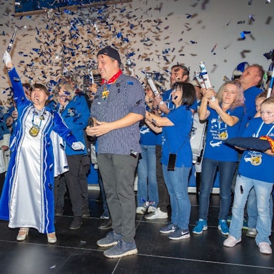 Anna Marian steht im blau-weißen Prinzessinnen-Orient auf einer Bühne und reckt jubelnd ein Zepter in die Höhe. Neben ihr steht Sacha Reichelt in grauer Hose und blau-weiß gestreiften Hemd. Im Hintergrund stehen viele Menschen in blauen T-Shirts, die Konfettikanonen in der Hand halten. Über alle regnet es silbernes und blaues Konfetti.