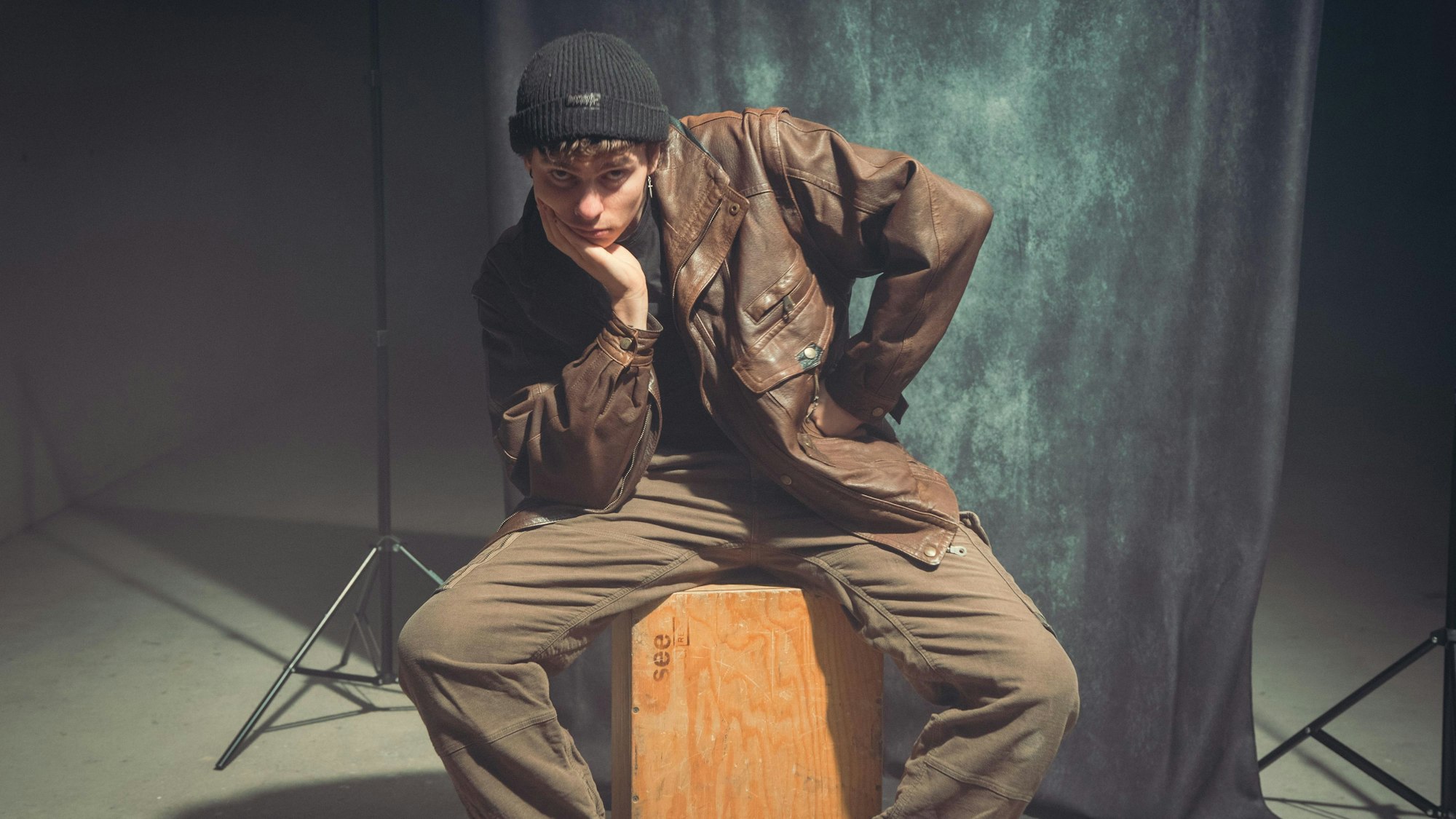 Ein junger Mann in Lederjacke und einer schwarzen Strickmütze sitzt auf einer Holzkiste in einem Fotostudio.