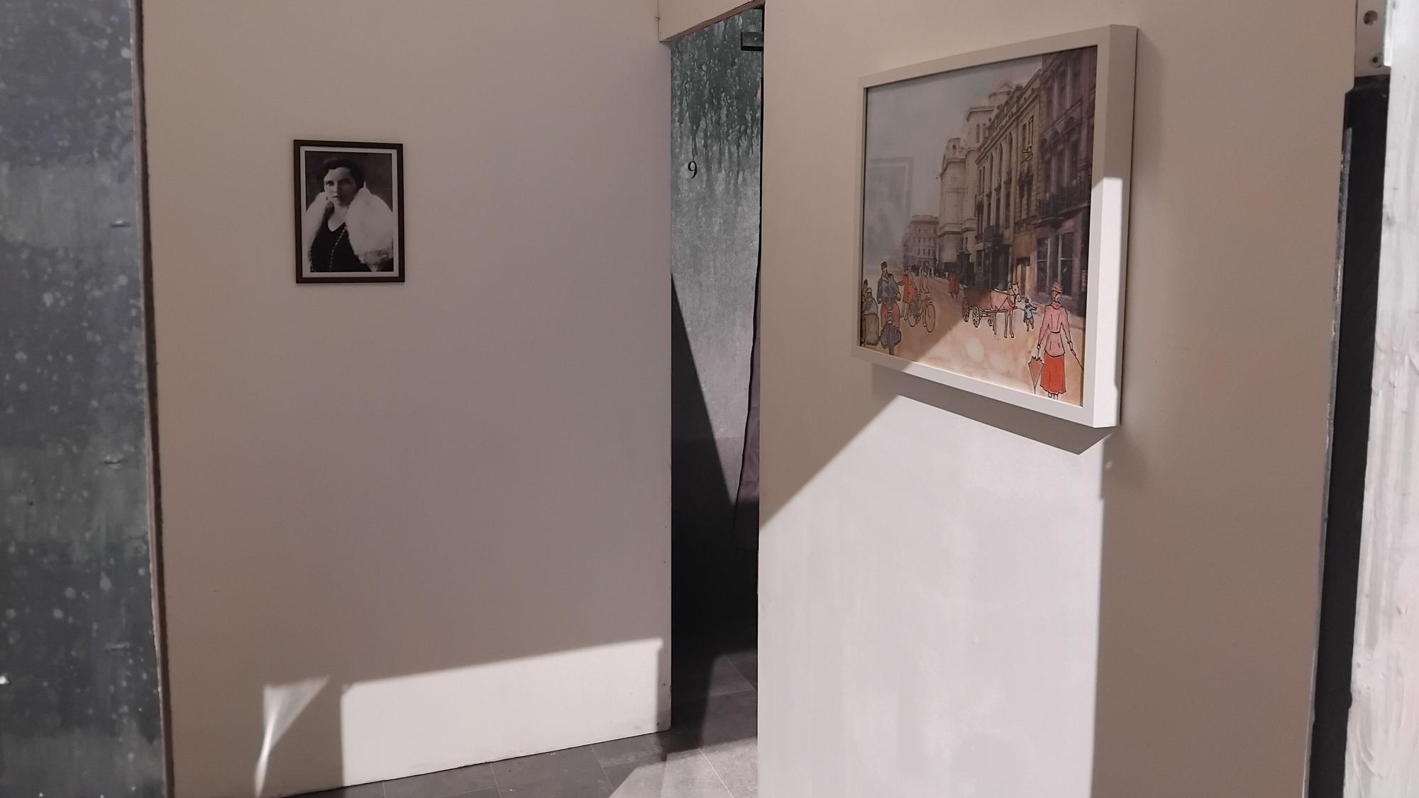 An einer weißen Wand hängen ein Schwarz-Weiß-Foto einer Frau und ein gezeichnetes Bild einer belebten Straße.