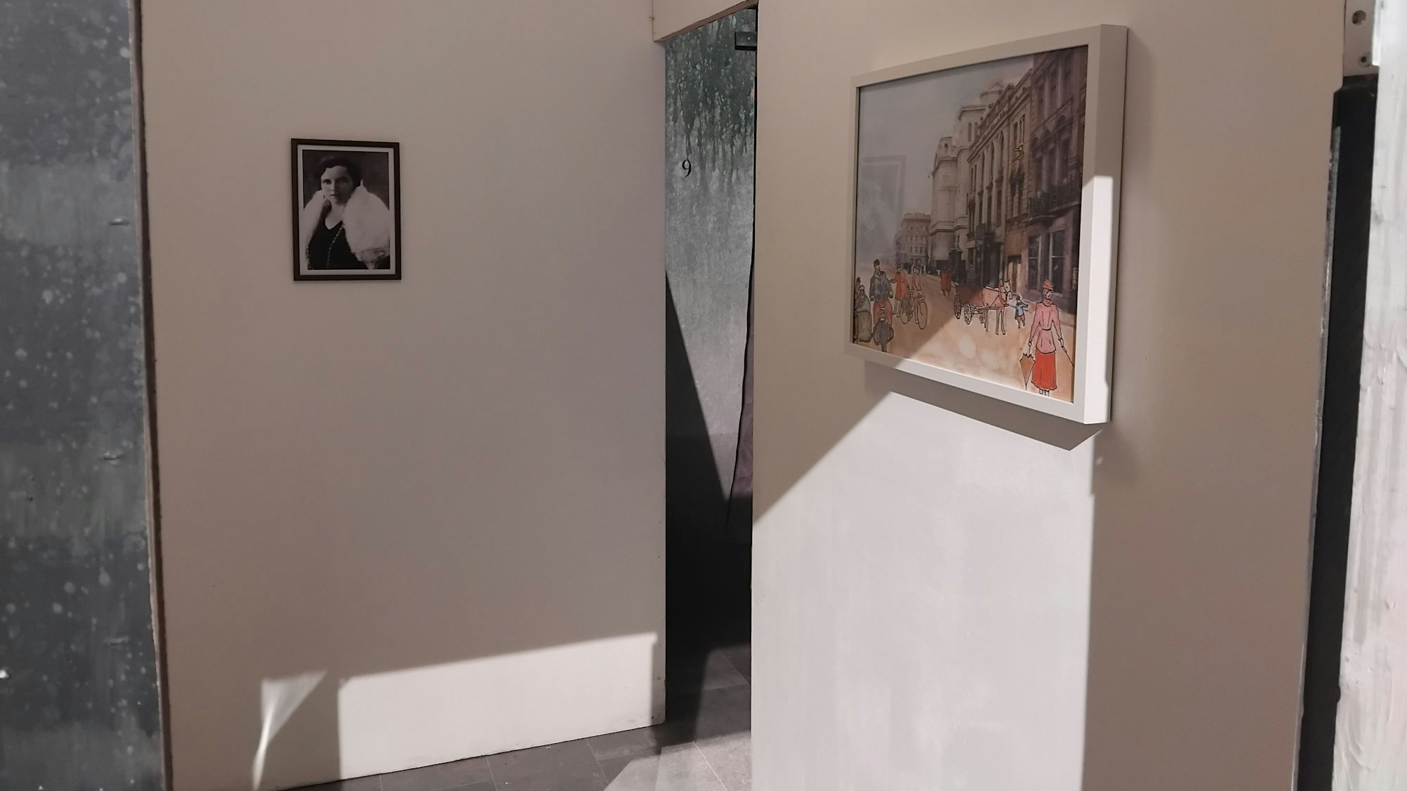 An einer weißen Wand hängen ein Schwarz-Weiß-Foto einer Frau und ein gezeichnetes Bild einer belebten Straße.